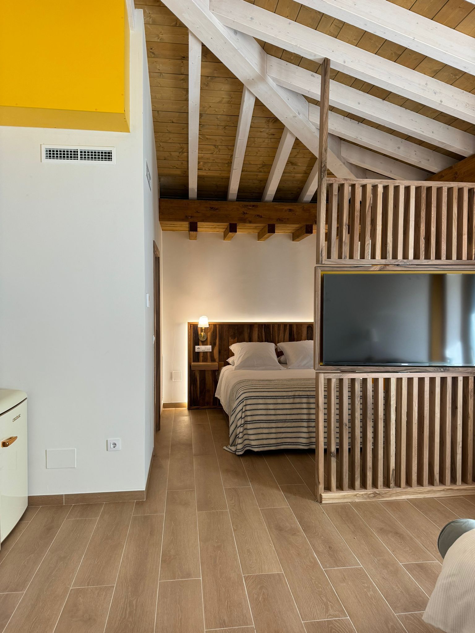 Dormitorio con detalles en madera. Cama visible tras un biombo de láminas y vigas de madera en el techo.