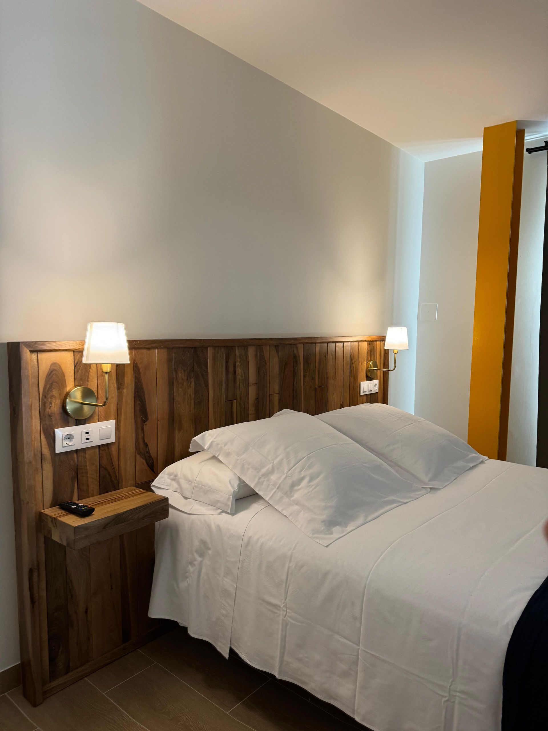 Dormitorio con cabecero de madera, ropa de cama blanca, lámparas de noche y una sección de pared de color naranja.