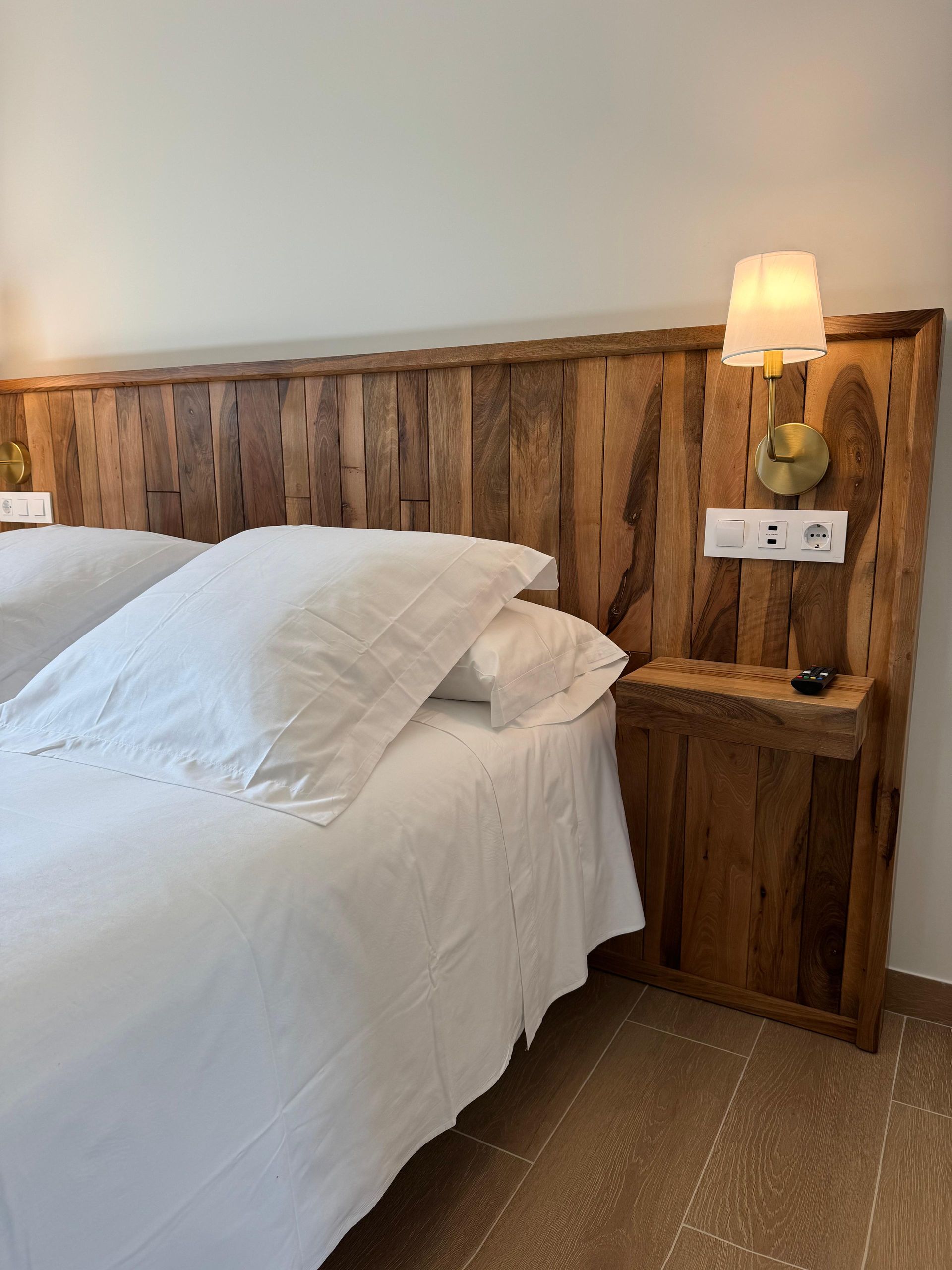 Cabecero de madera con mesita de noche, ropa de cama blanca, aplique de pared con pantalla y enchufes.