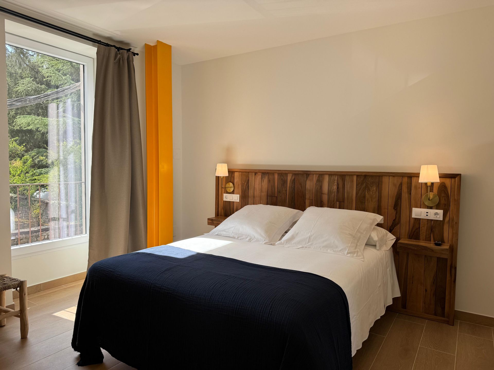 Dormitorio con cabecero de madera, ropa de cama blanca, manta azul y una ventana con cortinas amarillas y grises.