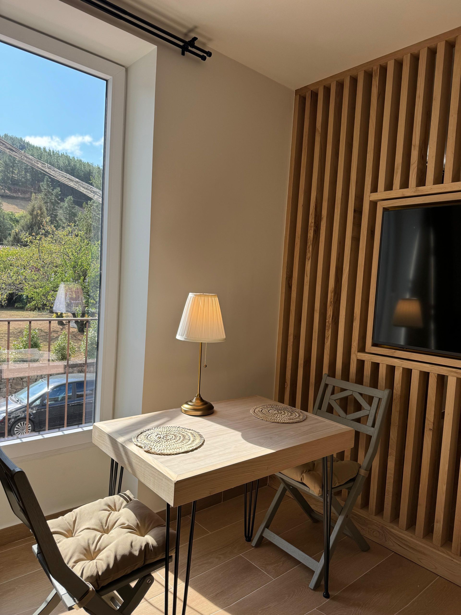 Un pequeño comedor con mesa, sillas, lámpara y televisor sobre una pared de listones de madera. vistas al exterior.
