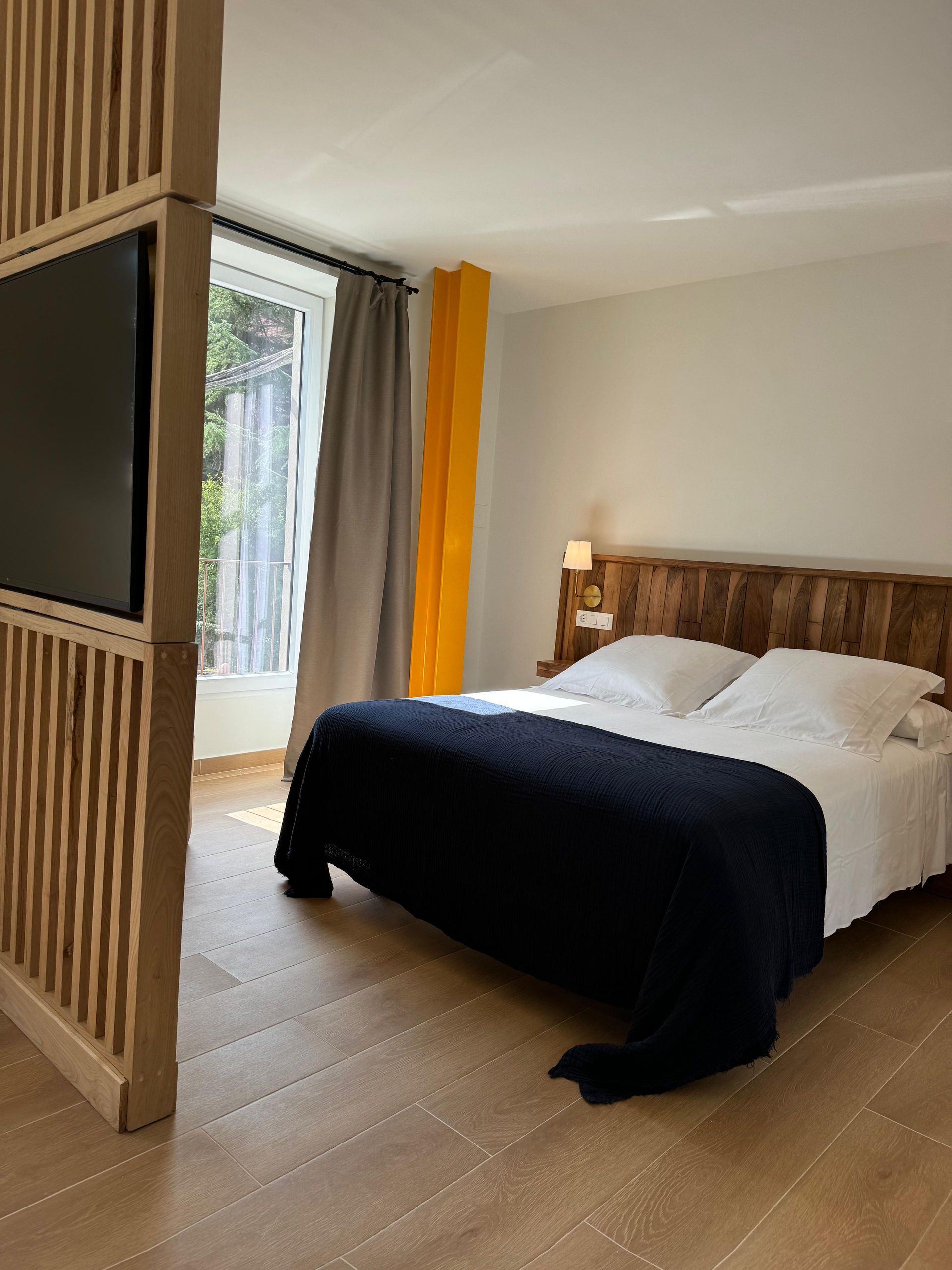Dormitorio con cabecero de madera, ropa de cama blanca y manta negra. Un biombo de madera con un televisor 