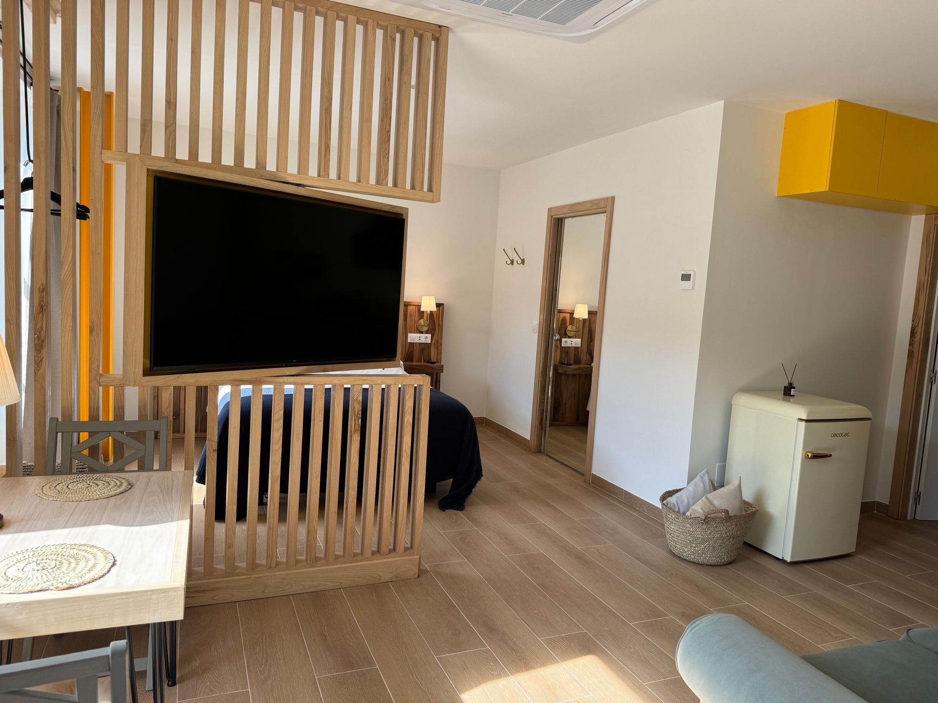 Acogedor estudio con mampara de madera, TV y mininevera. Madera clara y detalles en amarillo.