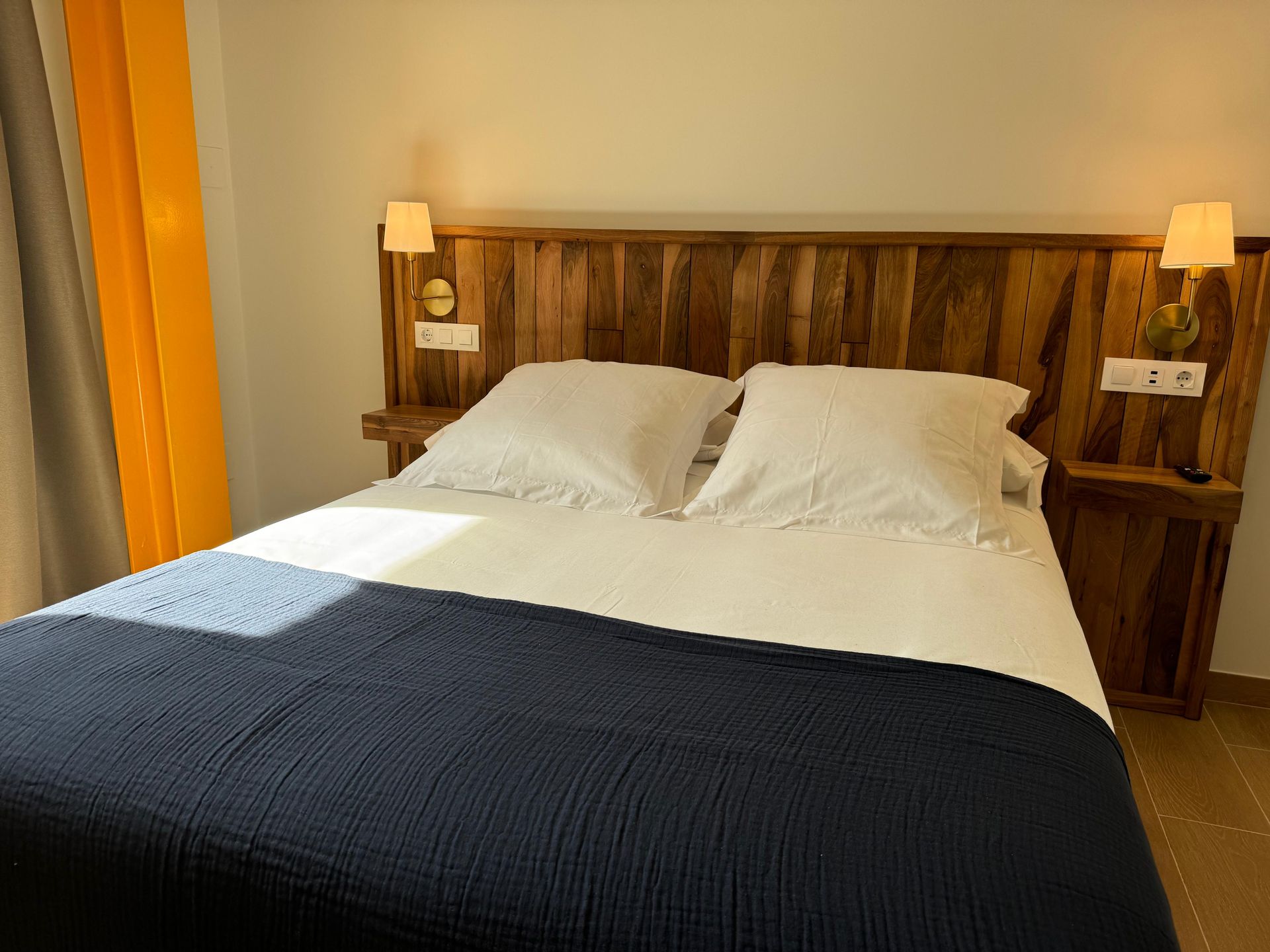 Dormitorio con cabecero de madera, ropa de cama blanca y una manta azul marino. Se ven cortinas amarillas.