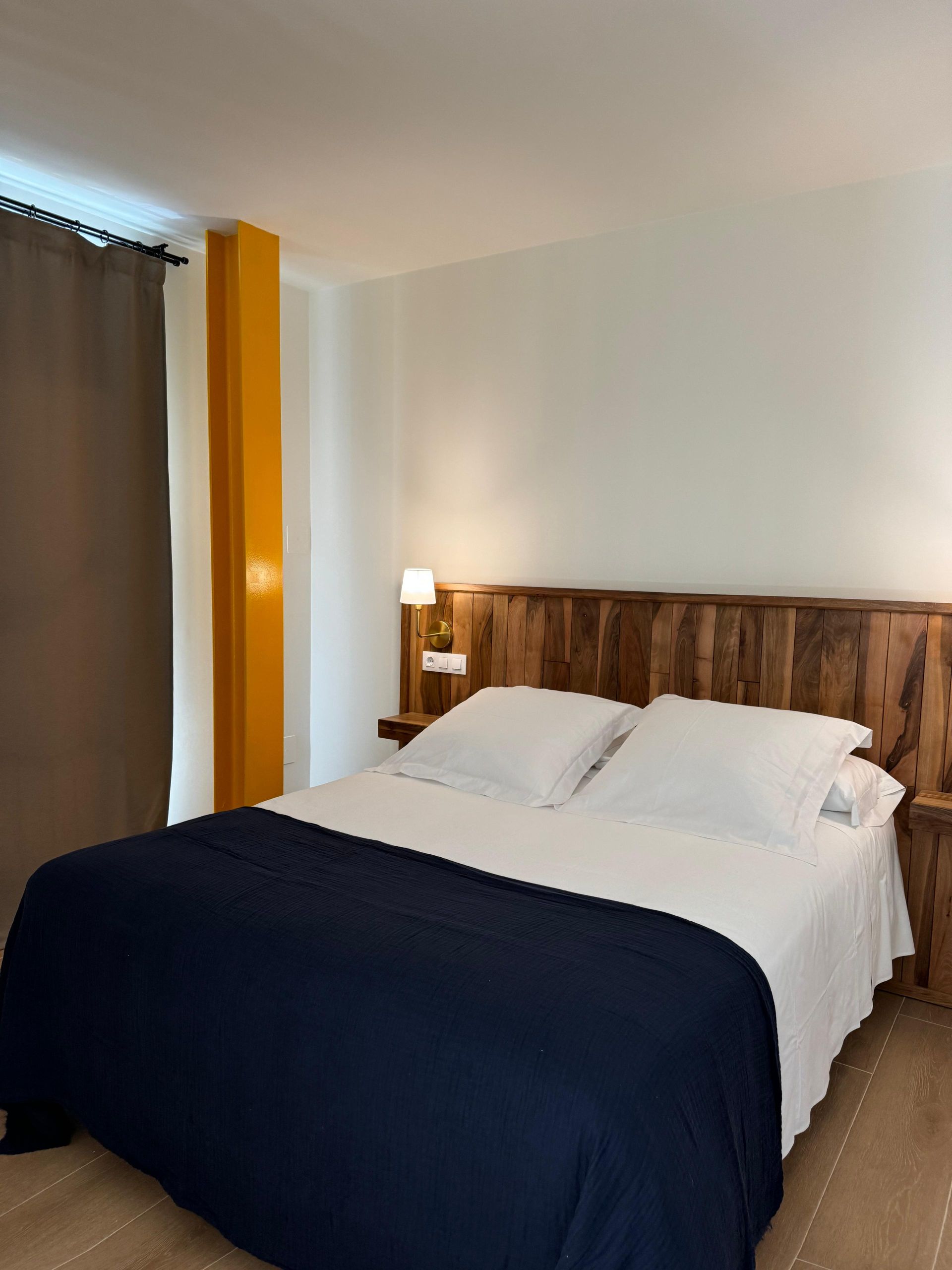 Dormitorio con cama, cabecero de madera, sábanas blancas, manta azul marino, pilar amarillo, cortinas marrones.