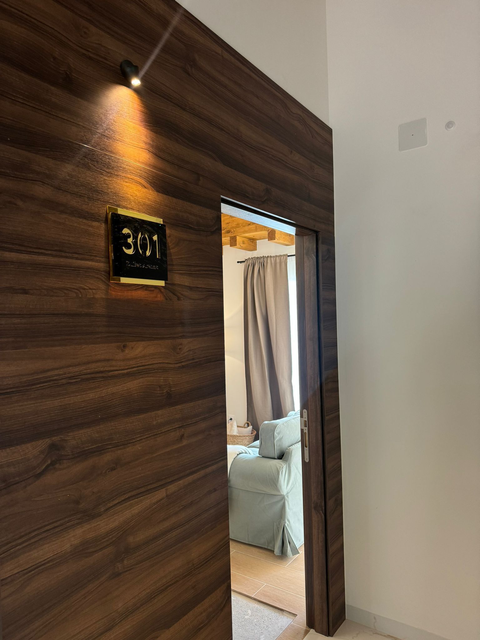 Puerta con paneles de madera y placa de dirección iluminada; vista a una habitación con un sofá.