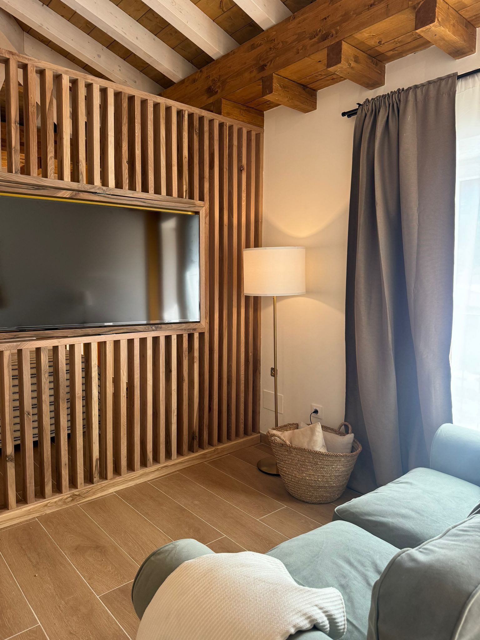 Sala de estar con listones de madera alrededor de un televisor. Alfombra beige, sofá gris, lámpara de pie. Vigas de madera.