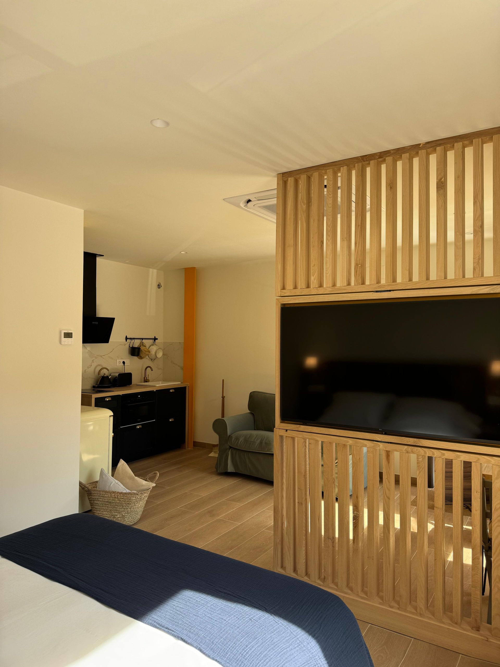Dormitorio con mampara de lamas de madera y TV, pequeña cocina al fondo.
