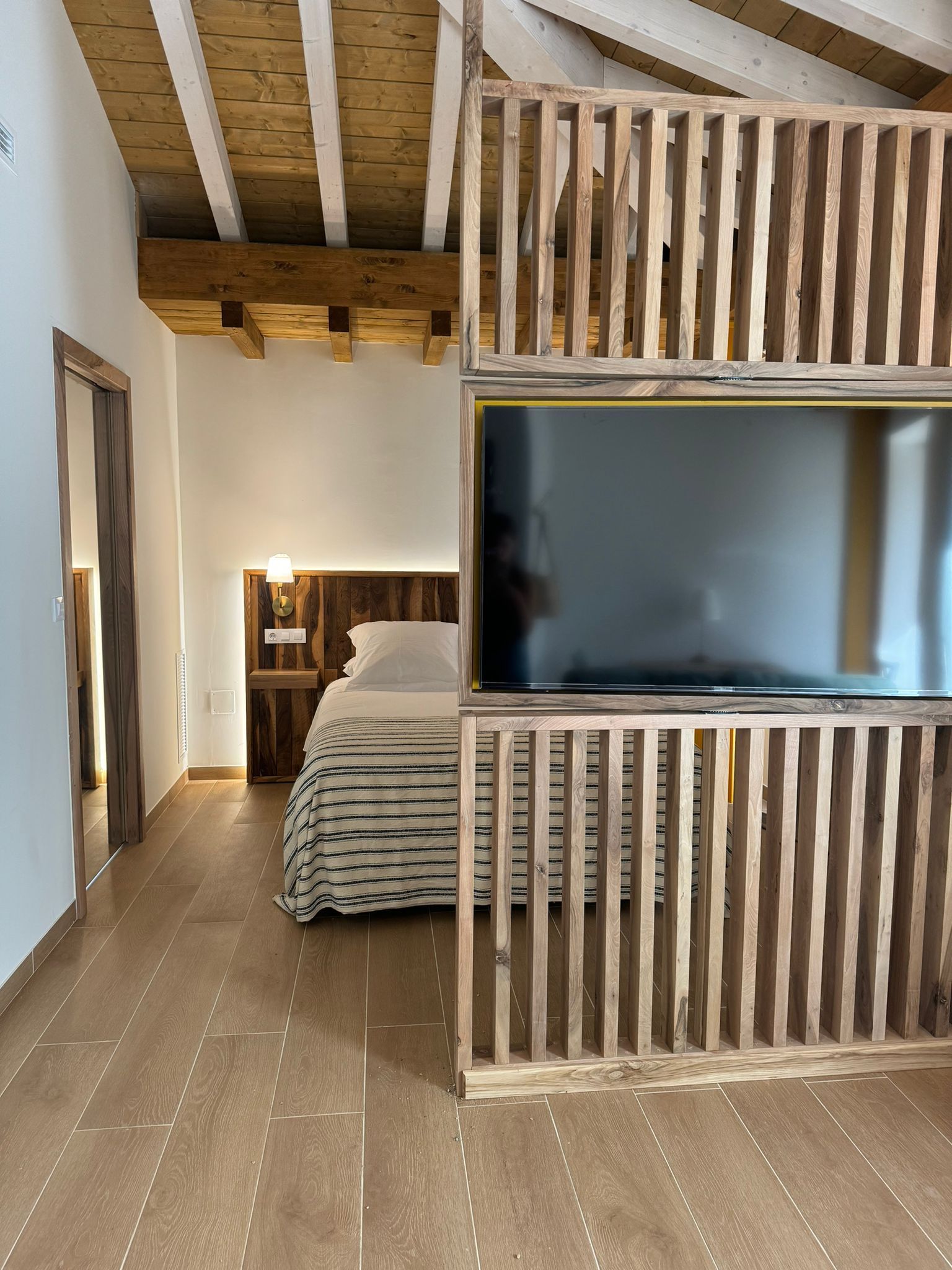 Dormitorio con biombo de listones de madera, cama con sábanas blancas y televisor. Suelo y techo de madera clara.