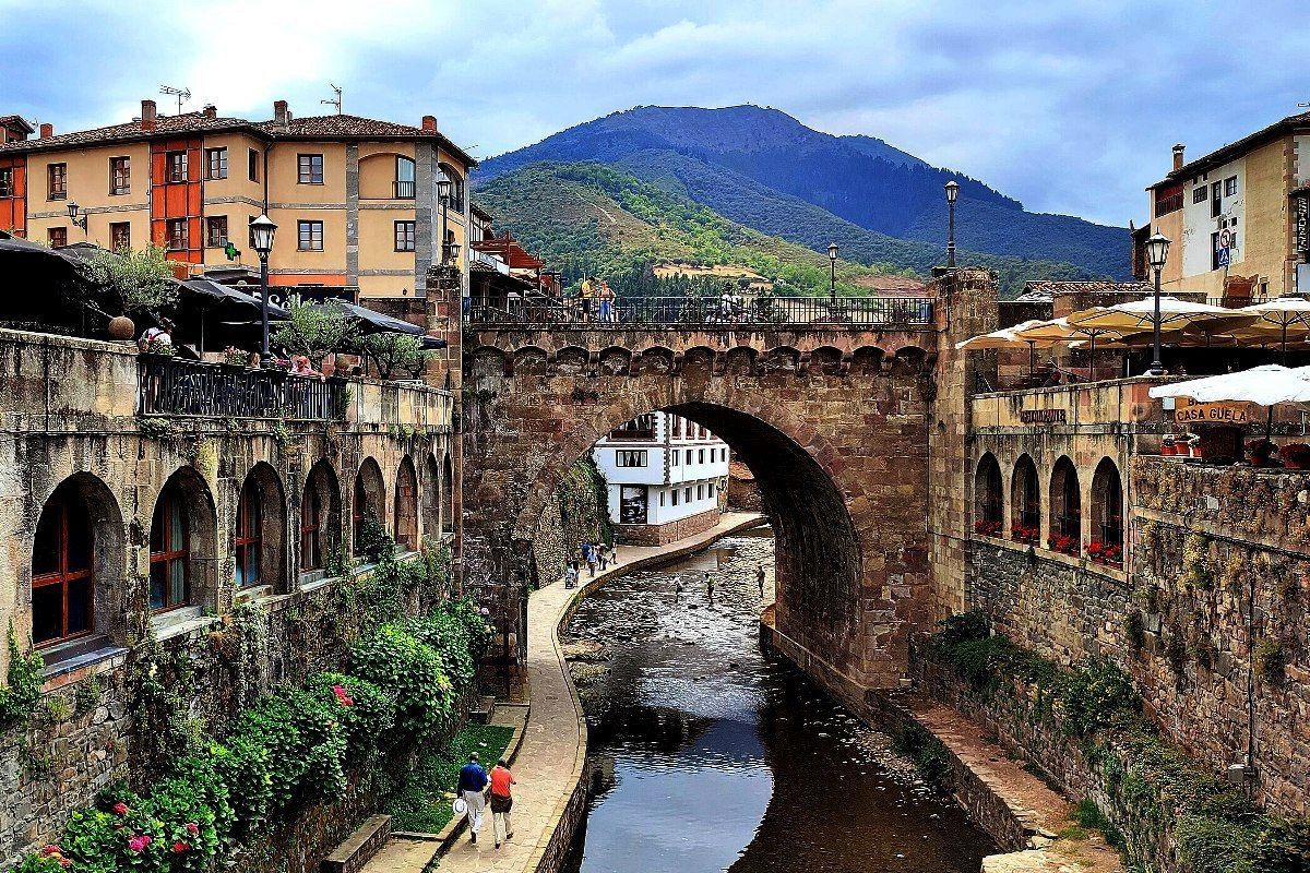 Puente de piedra sobre un río en un pueblo europeo con edificios coloridos y un fondo de montañas.