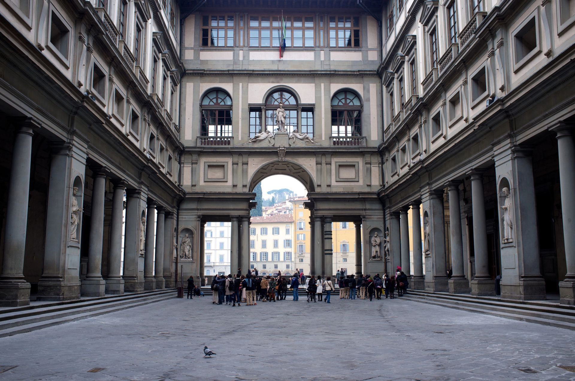 Il cortile della Galleria degli Uffizi a Firenze, con colonne in pietra, passaggi ad arco e un gruppo di visitatori in piedi al centro.