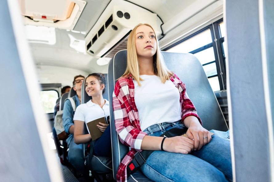 Gli studenti siedono in fila su un autobus, con uno studente concentrato in primo piano che indossa una camicia a quadri rossa.