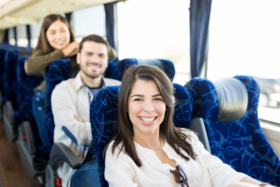 Tre persone sorridenti sedute in fila su un autobus con sedili a fantasia blu.