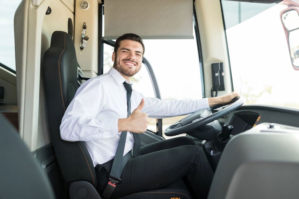 Un autista professionista sorridente, in camicia bianca e cravatta, fa il segno del pollice in su mentre è seduto al posto di guida di un autobus.