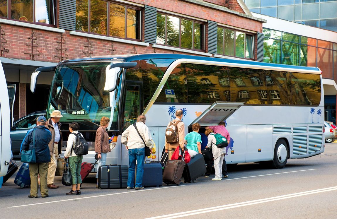 Un gruppo di persone carica i bagagli nel vano laterale di un autobus bianco parcheggiato davanti a un edificio in mattoni.