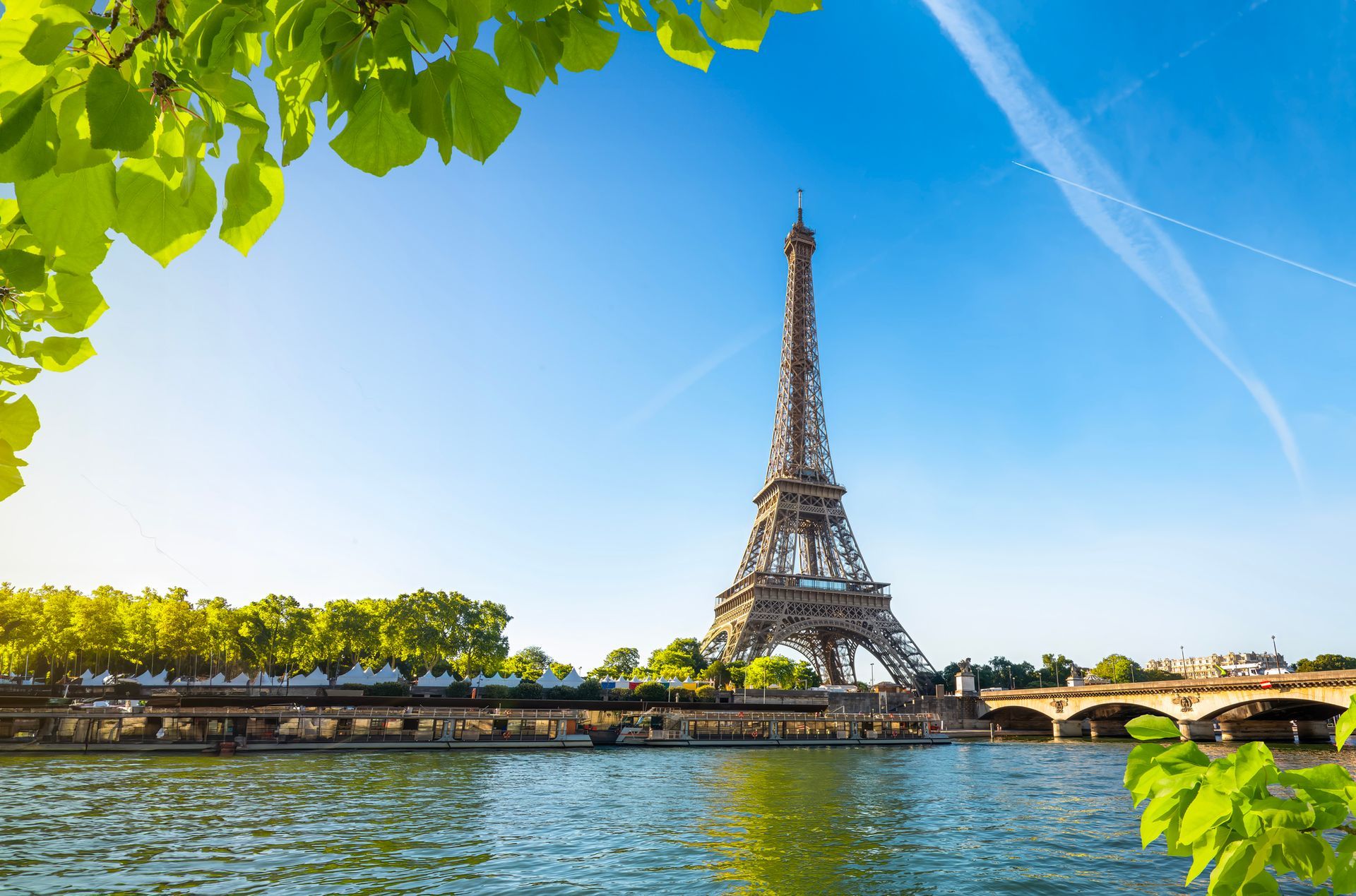 Sotto un cielo azzurro brillante, la Torre Eiffel si erge maestosa sulle rive della Senna a Parigi, incorniciata dal verde delle foglie.