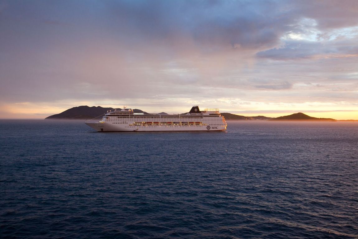 Al tramonto, una grande nave da crociera galleggia su un mare calmo e scuro, stagliandosi contro un cielo nuvoloso e isole lontane.