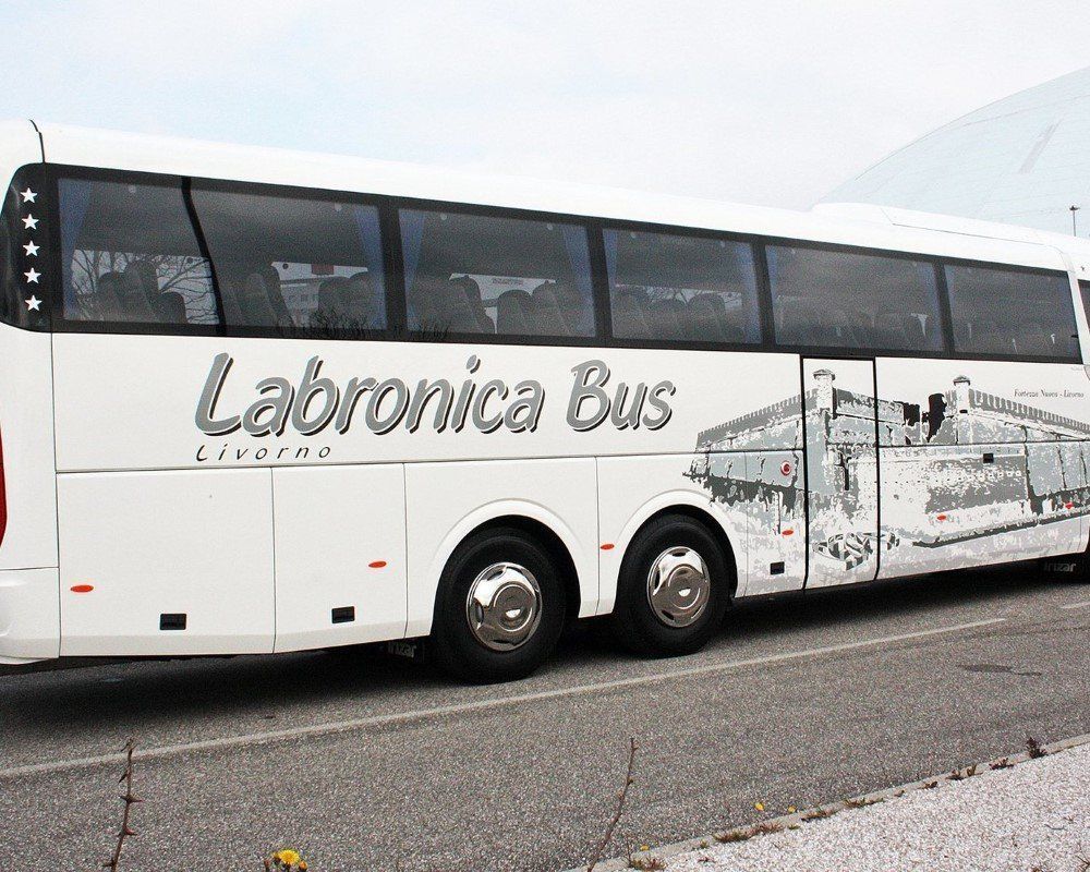 Un pullman turistico bianco della compagnia Labronica Bus è parcheggiato a lato della strada, con una grafica panoramica raffigurante un edificio sul fianco.