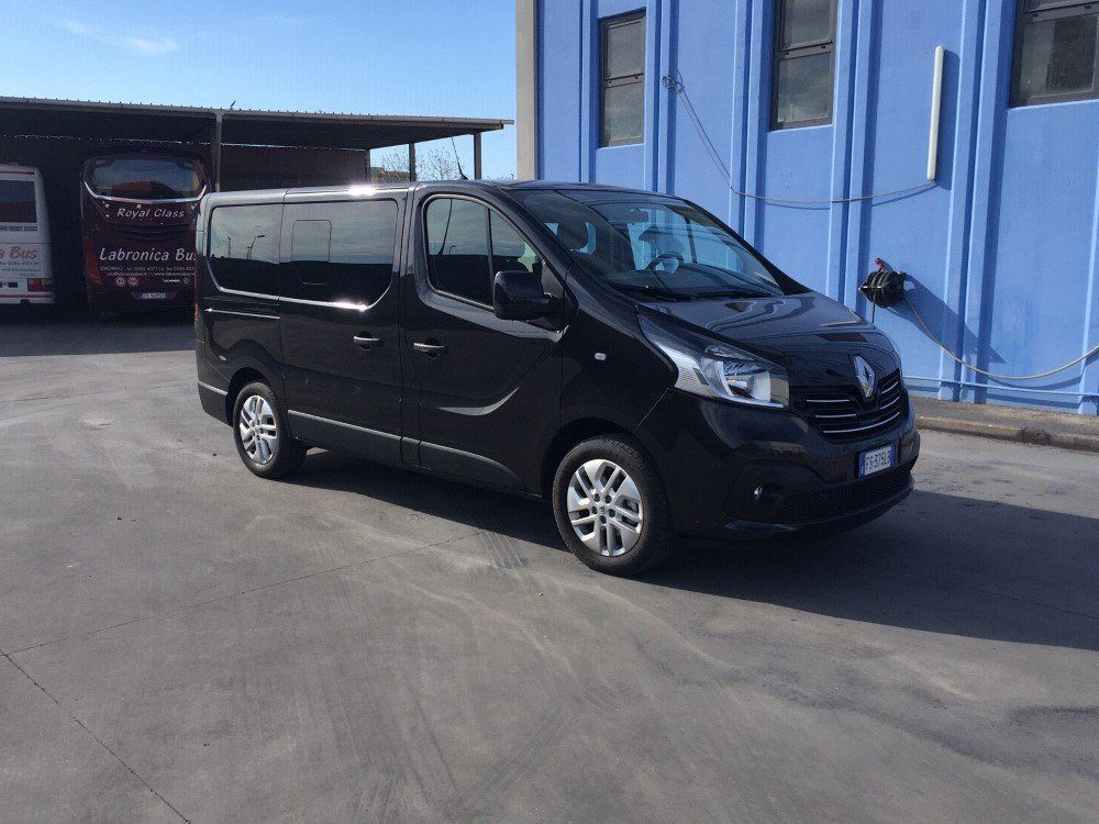 Un furgone passeggeri Renault Trafic nero parcheggiato su un piazzale asfaltato di fronte a un edificio industriale blu.