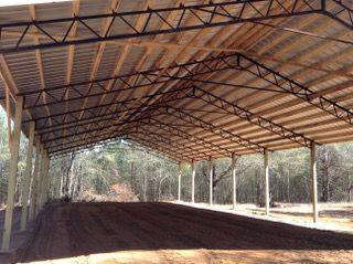 A large metal structure with a wooden roof is surrounded by dirt and trees.