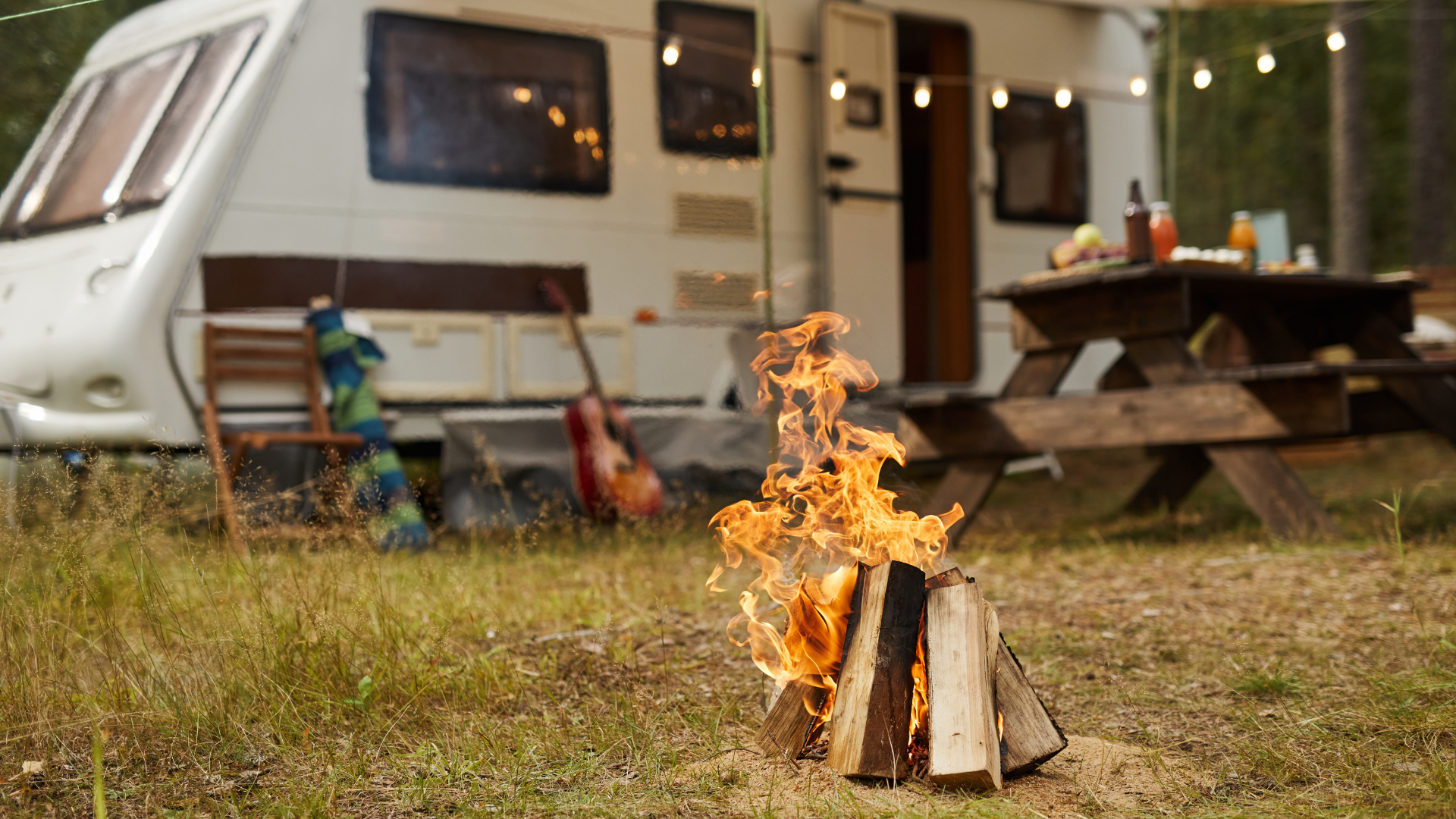 Campfire burning beside a camper and picnic table at an outdoor campsite at dusk