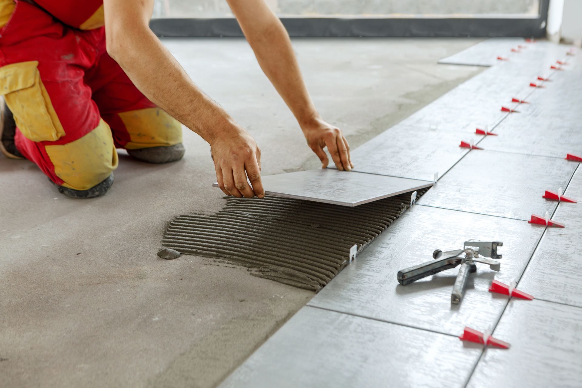 Worker spreading tile adhesive on a concrete floor with a trowel, alongside tile spacers and tools