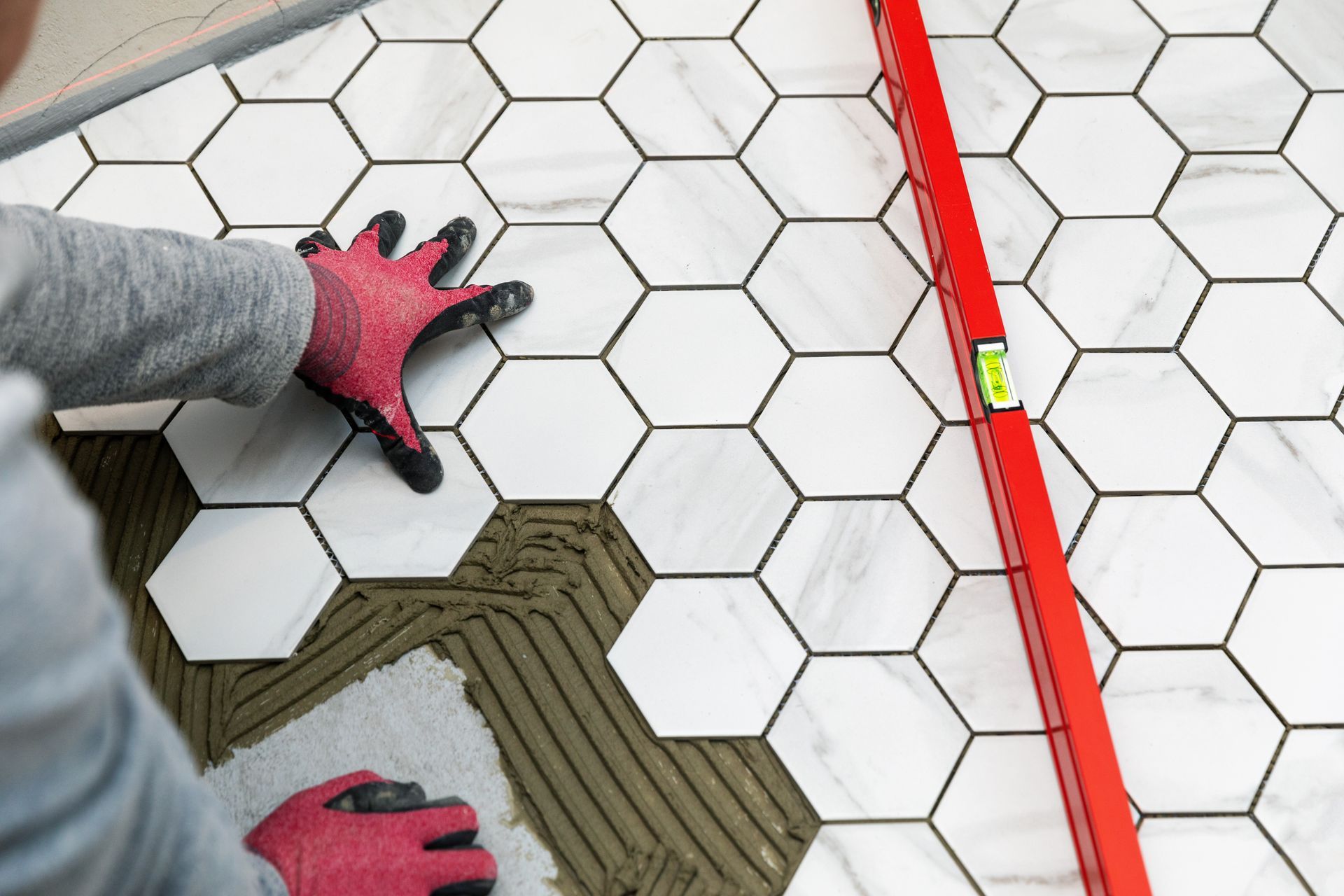 Hands in red gloves aligning hexagonal floor tiles beside a red straightedge ruler.