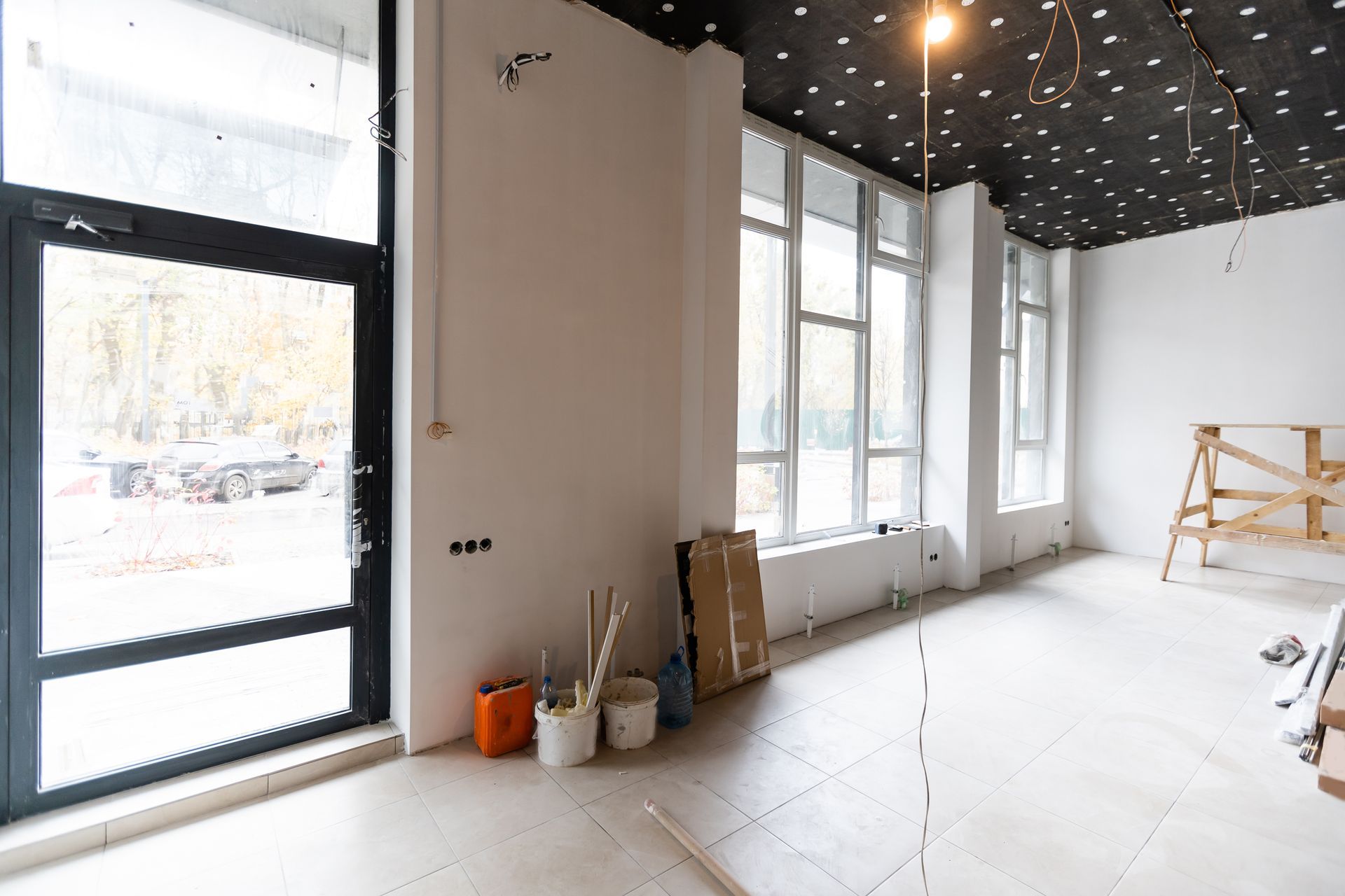 Bright unfinished interior with large windows, white walls, exposed ceiling, and painting supplies on the floor