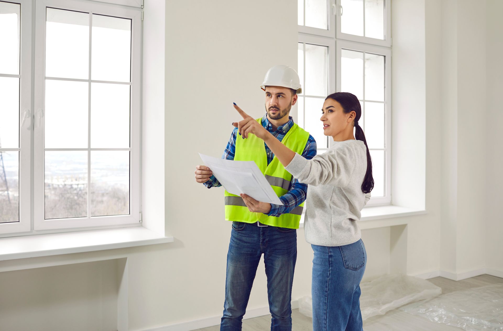 Two construction workers in hard hats review blueprints at a building site with excavator nearby