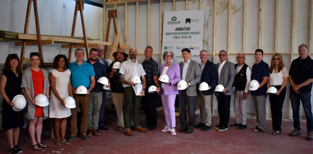 Group of people holding white hard hats posing in front of a wall under construction.