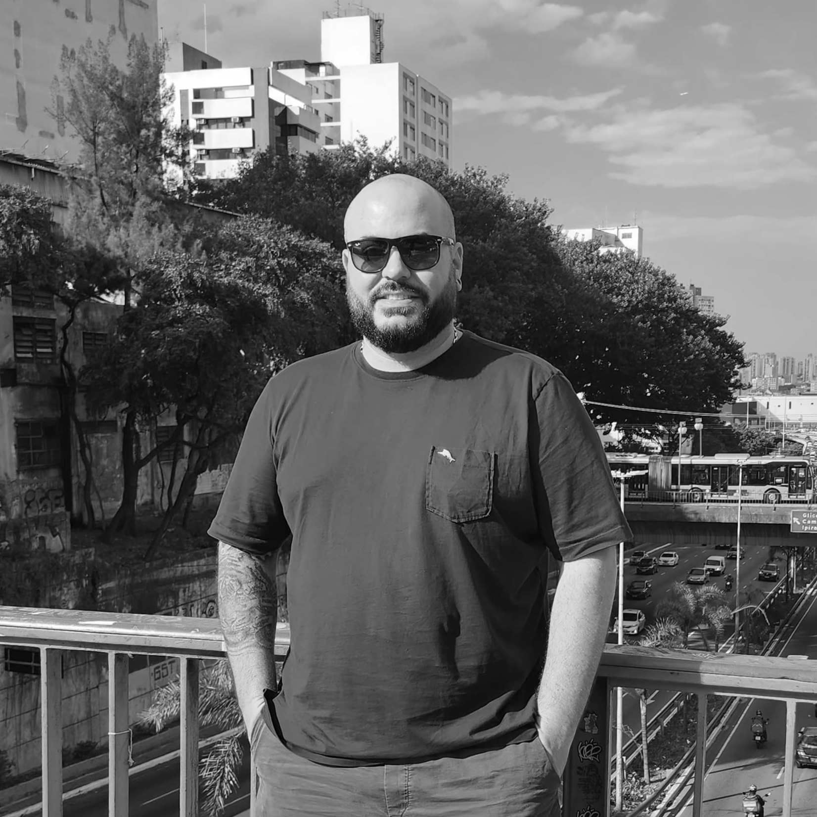 Un hombre con barba y gafas de sol está parado en un puente frente a una ciudad.