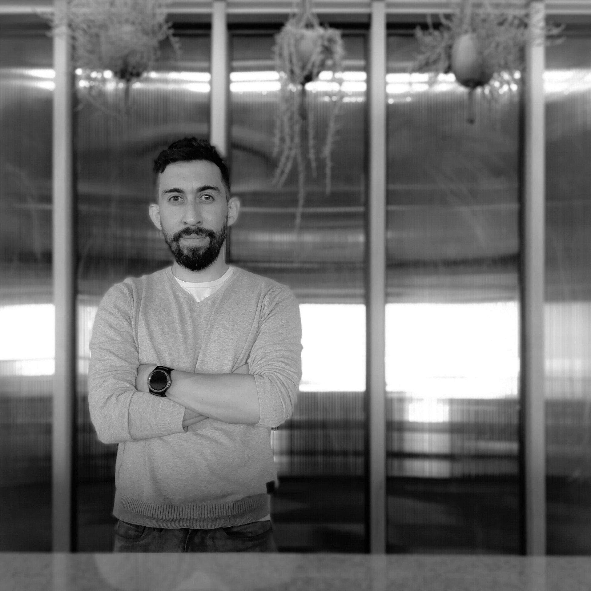 Un hombre con barba está parado frente a una ventana con los brazos cruzados.