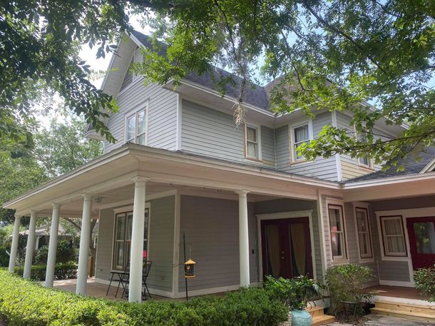 Two-story house with a large porch and light gray siding, partially obscured by green trees.