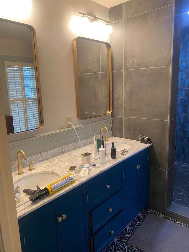 Bathroom with blue vanity, marble countertop, gold fixtures, mirrors, and gray tiled walls.