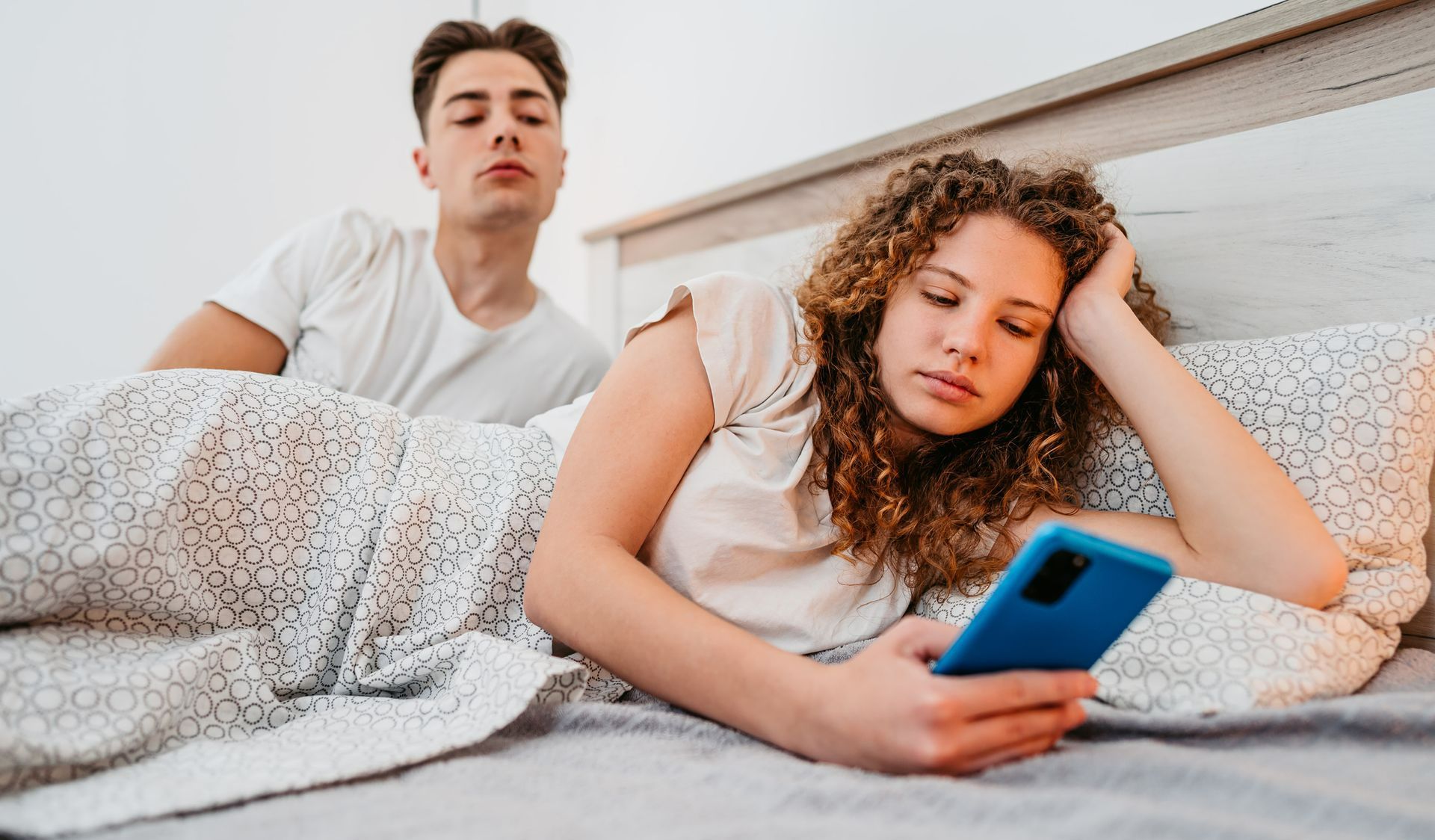 A woman is laying in bed looking at her phone while a man looks on.