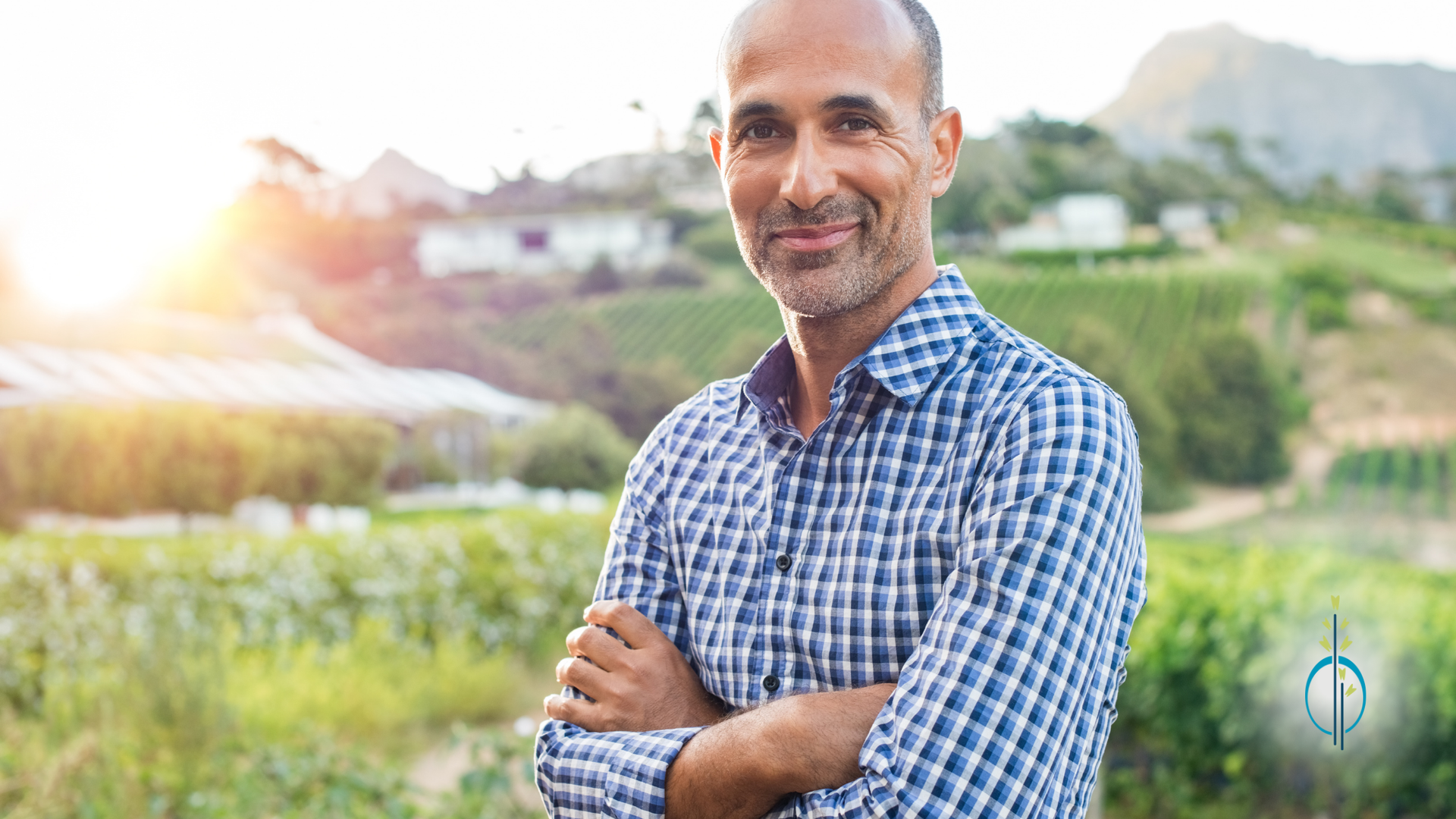 A man in a plaid shirt is standing in a field with his arms crossed.