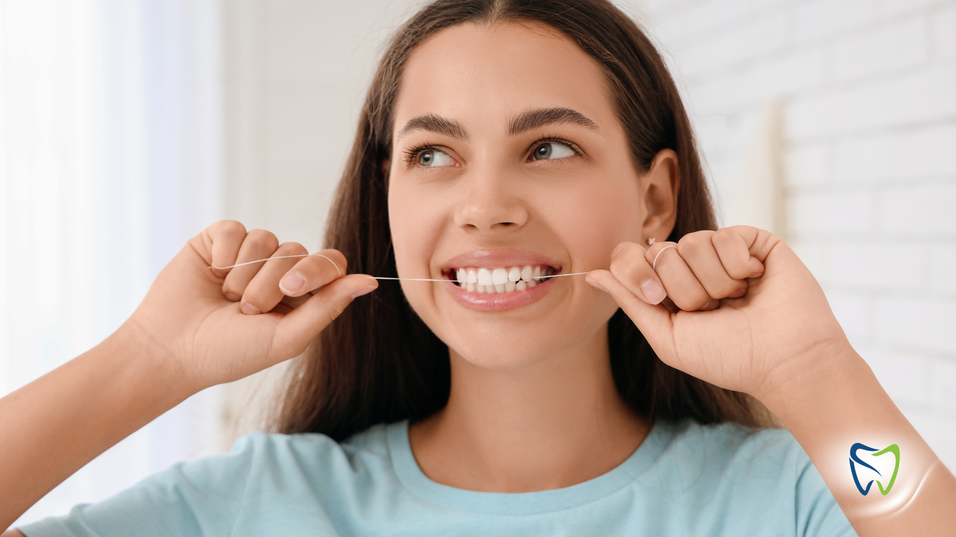 Woman flossing teeth indoors, smiling, wearing a blue shirt.