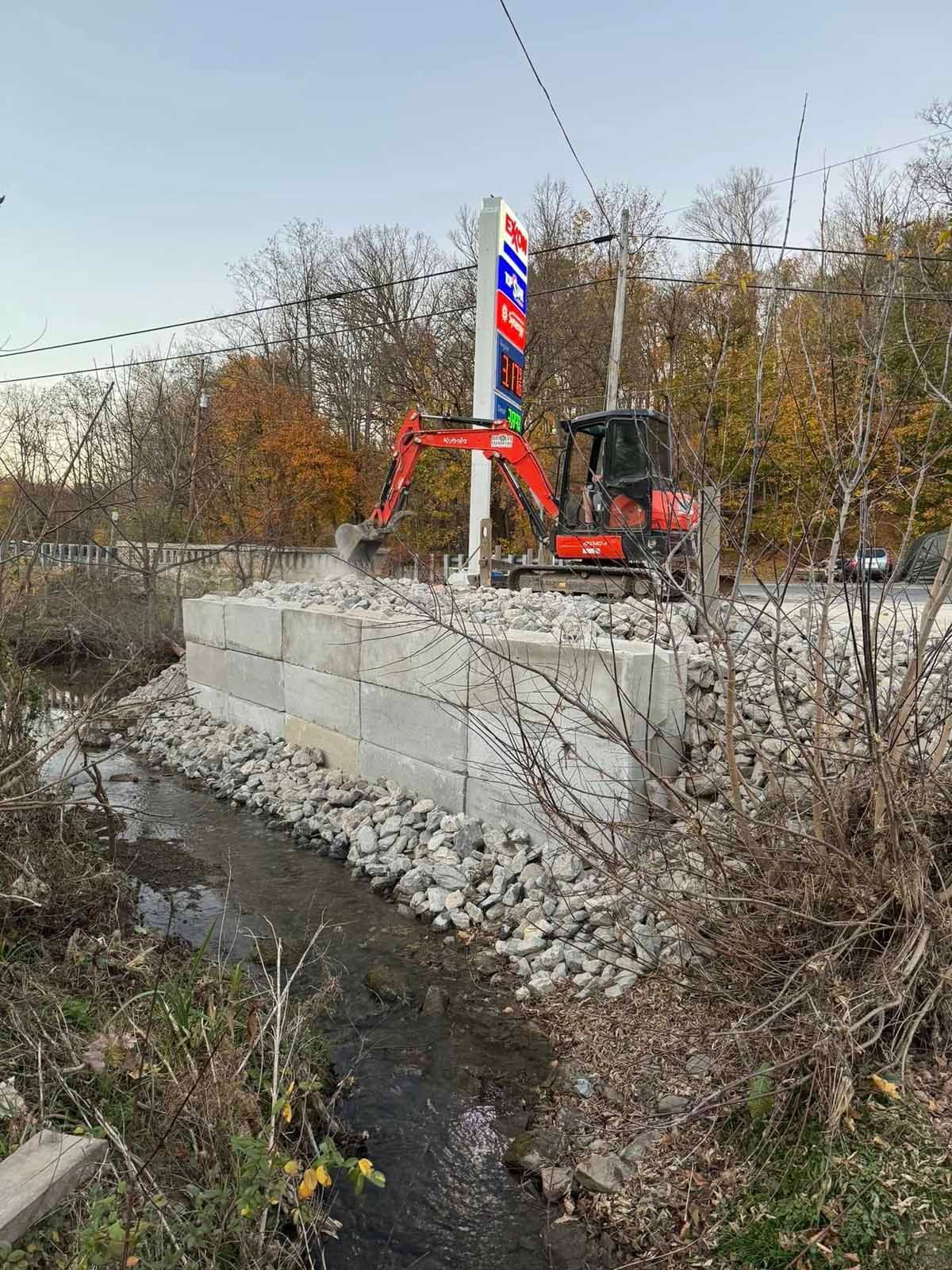 A small excavator is working on a bridge over a stream.