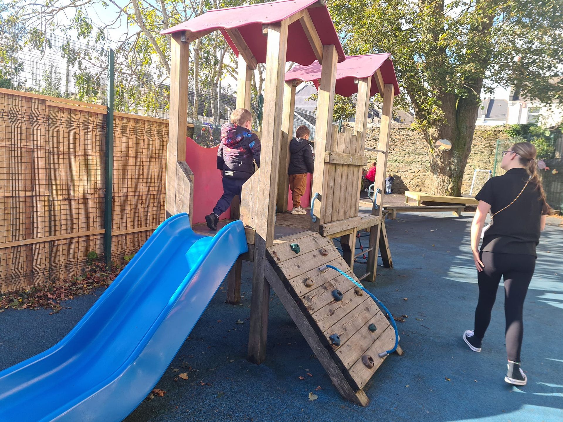 A child is playing on a slide at a playground while a woman walks by.