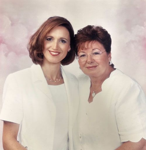 Two women in white outfits smile, posing together. Pink and white background.
