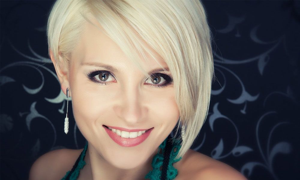 Woman with short blonde hair smiles, wearing dangling earrings, against a floral background.