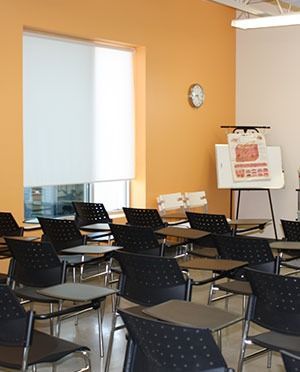 Classroom with rows of desks, orange and beige walls, clock, window with white shade, whiteboard.