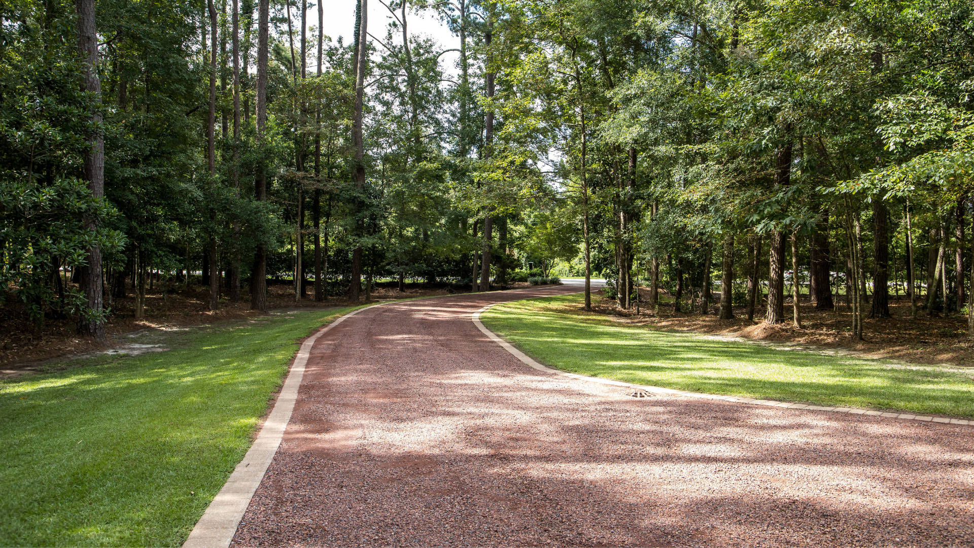 A curved gravel driveway with a stone border winds through a lush, sunlit forest landscape.