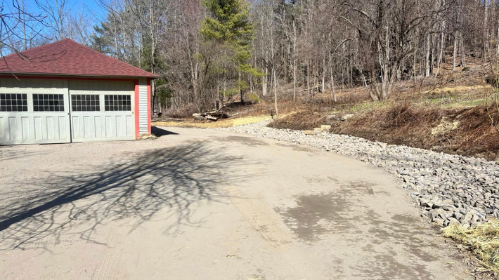 A cream-colored garage with a red roof stands next to a gravel driveway bordered by stones and a forest in the background.