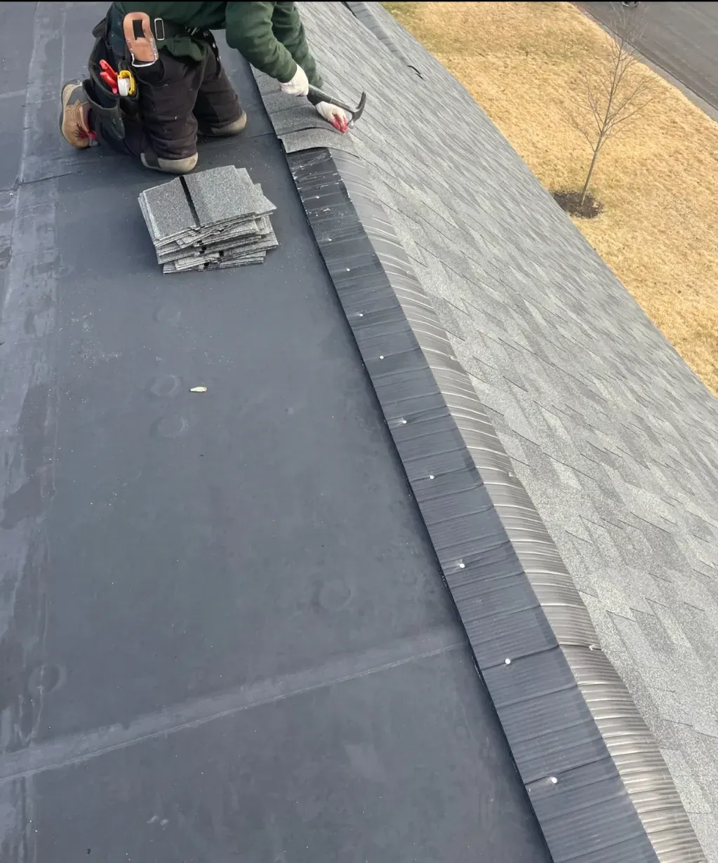 A roofer on a sloped roof installs shingles. He kneels near a stack of shingles, applying sealant.