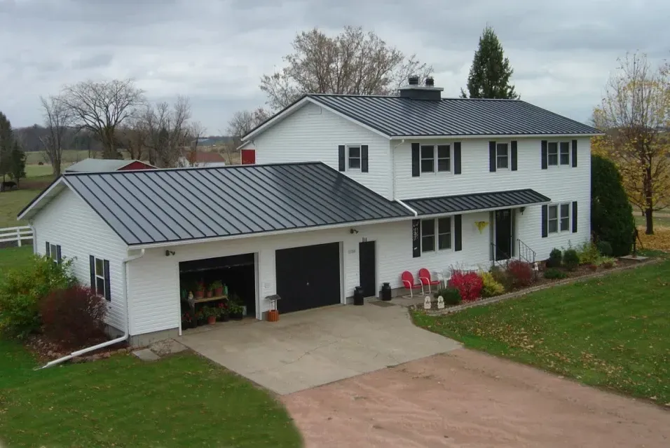White two-story house with a black roof and attached garage. Red chairs sit on the porch; lawn surrounds a concrete driveway.