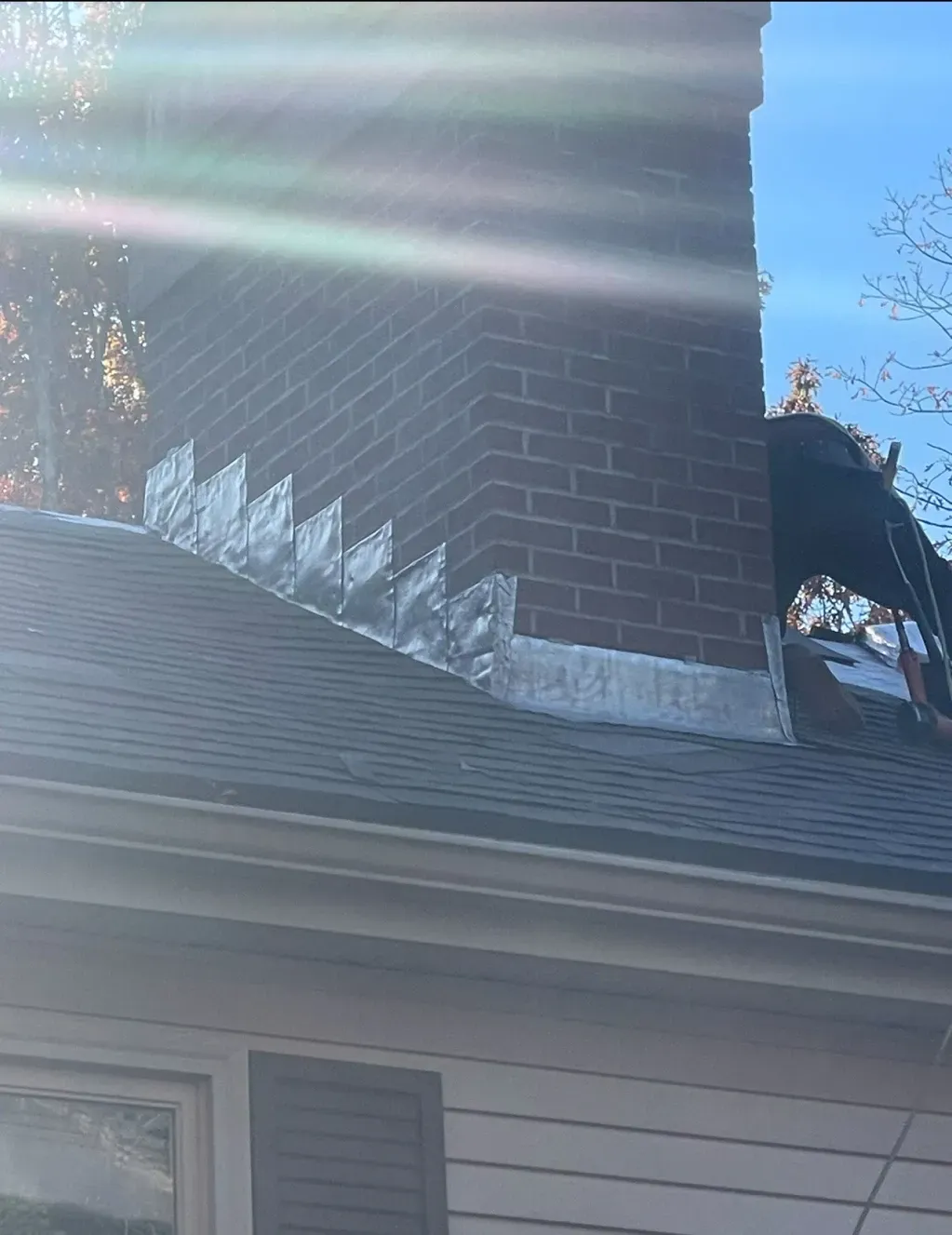 Brick chimney on a gray-shingled roof with flashing, in a sunny outdoor setting. A person is visible on the roof.