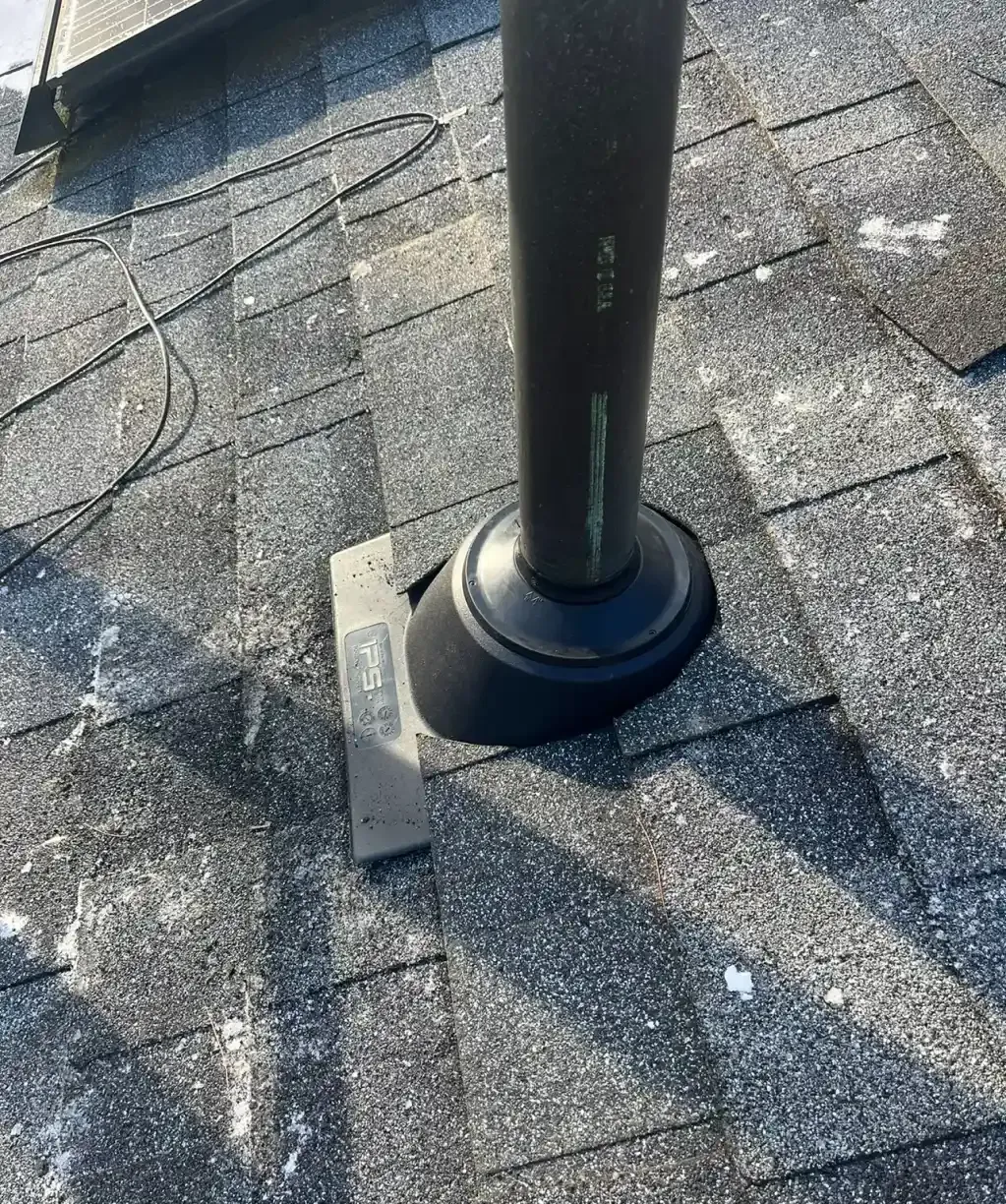 Black pipe vent protruding from a gray shingled roof with a black base and silver flashing. There are small patches of snow on the roof.