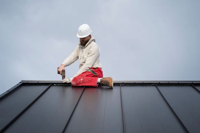 A man is kneeling on a roof using a drill.