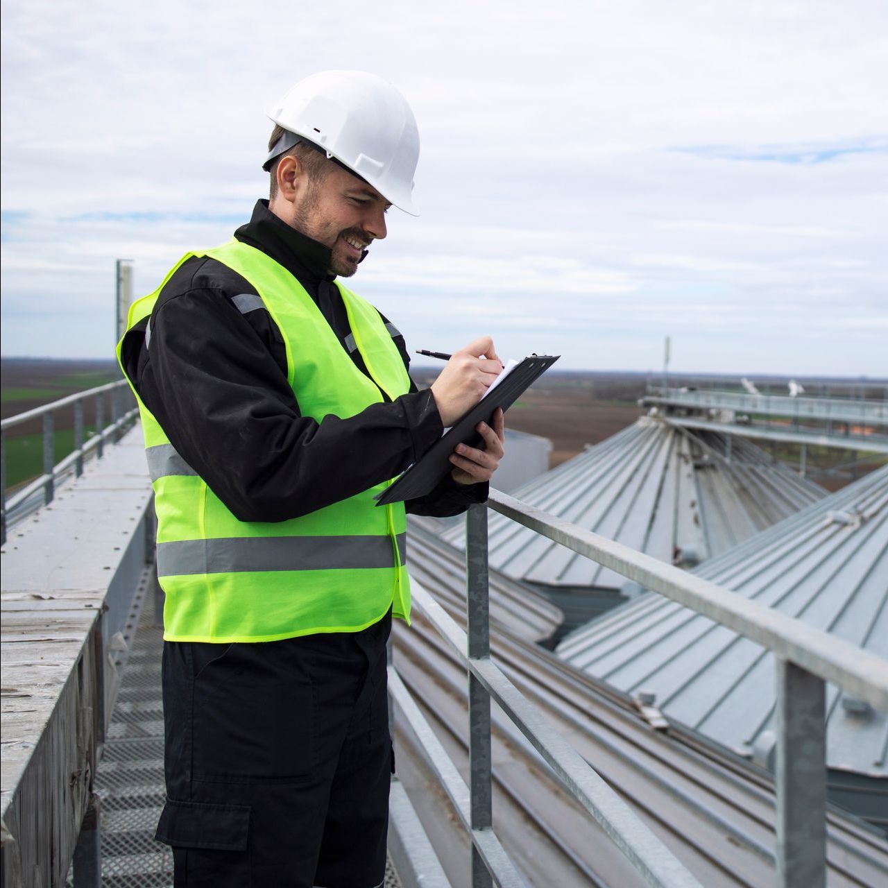 A man in a hard hat and safety vest is standing on a balcony looking at a tablet.